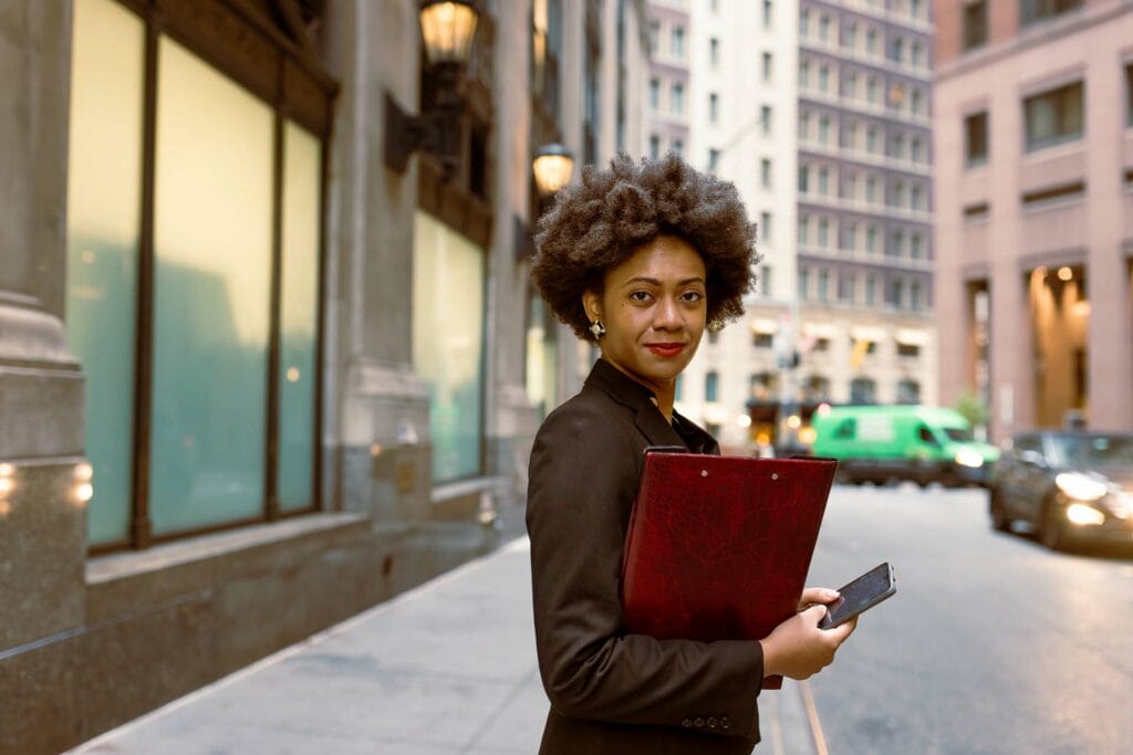 A process server with curly hair stands on a city sidewalk, holding a red folder and a smartphone, and wearing a black jacket. Buildings and cars are in the background.